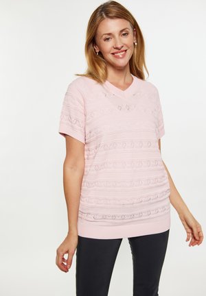 Lichtroze gebreide top met korte mouwen en een V-hals, met een textuurpatroon en horizontale details. Gedragen met donkere broek.
