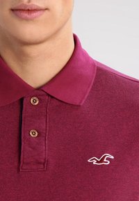 Burgundy polotröja i strukturerad tyg, med klassisk krage, tre knappar och en vit broderad logotyp på bröstet.