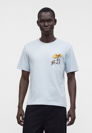 Homme portant un t-shirt bleu clair avec un petit motif représentant une voiture jaune et quatre personnes près de la poitrine gauche, debout devant un fond uni.