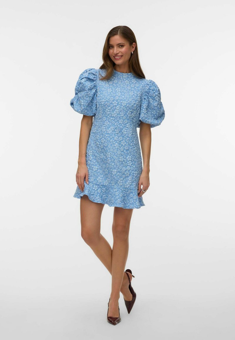 Femeie în picioare, purtând o rochie scurtă albastră cu imprimeu floral, mâneci bufante și tiv cu volane, asortată cu pantofi stiletto de culoare închisă, cu vârf ascuțit.