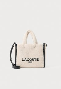 Velika kremasta torba od lažnog janjećeg krzna s crnim detaljima, gornjim ručkama i odvojivim remenom za rame. Ima izvezeno "LACOSTE 1933".