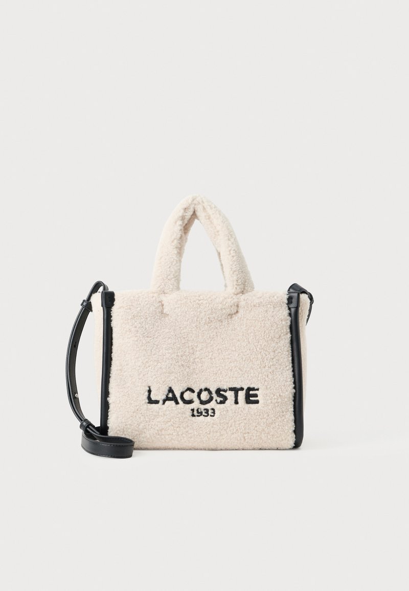Velika kremasta torba od lažnog janjećeg krzna s crnim detaljima, gornjim ručkama i odvojivim remenom za rame. Ima izvezeno "LACOSTE 1933".