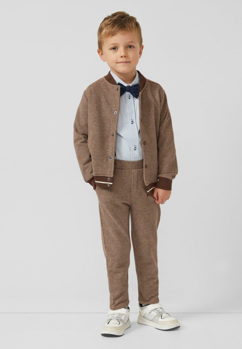 Brązowy sweter z fakturą, jasnoniebieska koszula, ciemna mucha, pasujące brązowe spodnie, białe trampki z szarymi akcentami, wszystko zaprojektowane dla dzieci.