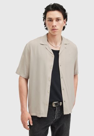 Chemise à manches courtes, de couleur beige clair, en tissu lisse, avec un col retroussé et des boutons beiges, associée à un débardeur noir.