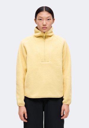 Jeune personne aux cheveux noirs et lisses porte un pull polaire jaune clair à col montant avec une demi-fermeture éclair, associé à un pantalon noir.