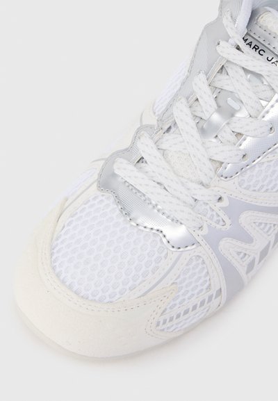 Fehér sportcipő, hálós és bőr anyagokból. Ezüst díszítésekkel, texturált mintákkal és lapos fehér fűzőkkel rendelkezik. Lekerekített orrformával.