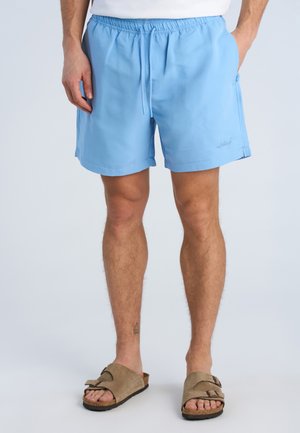 Mand iført lyseblå knælange shorts med snøre i taljen og brune ruskindssandaler, stående med hænderne i lommerne.