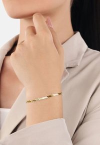 Guldtonad armband med en vriden design, som har en glänsande, reflekterande yta. Bärs på en hand mot en ljus tygbakgrund.