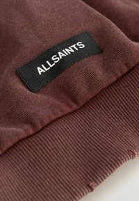 Maroon dukserica s rebrastim rubom i crnom etiketom na kojoj piše "ALLSAINTS" bijelim slovima. Glatka tekstura, ležeran dizajn.