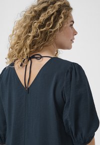 Blusa color azul marino con un acabado texturizado, que presenta un diseño de espalda en V y un cierre con lazo. Tiene mangas cortas abullonadas y un corte relajado.
