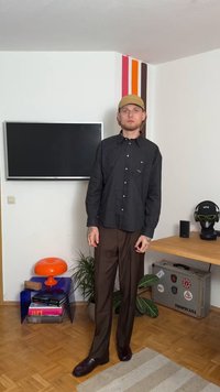 Chemise à rayures noires et blanches, pantalon marron, chaussures en cuir marron, casquette jaune. Le décor comprend des plantes et un fond de télévision.