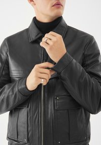 Giacca in pelle nera con un design strutturato, chiusura con zip e due tasche anteriori. Presenta un colletto classico e una texture liscia.
