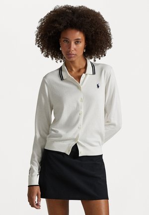 Cream cardigan with a black striped collar, front buttons, and a small logo. Paired with a black skirt, featuring a fitted, smooth texture.
