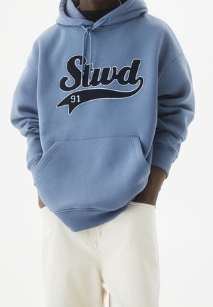 Persoon die een lichtblauwe hoodie draagt met de tekst "Stud 91" op de voorkant en gebroken witte broek, handen in de zakken van de hoodie, neutrale achtergrond.