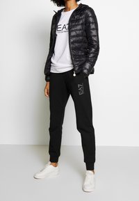 Svart vadderad jacka över en vit t-shirt med logotyp, kombinerat med svarta sweatpants och vita sneakers. Slät textur, avslappnad design.