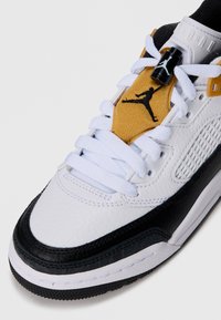 Baltas ādas sneakers ar melniem akcentiem, auduma mēle ar dzeltenu ielāpu un melnu Jumpman logo, plakani balti šņorējumi, teksturēta virsma.