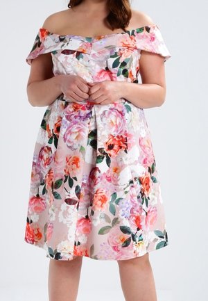 Woman wearing off-shoulder, knee-length dress with pink, orange, white, and purple floral print, hands clasped at waist.