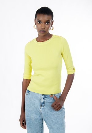 Donna con capelli corti che indossa orecchini a cerchio dorati, maglietta gialla con maniche a tre quarti e bottoni, e jeans azzurri chiari, in piedi contro uno sfondo bianco.