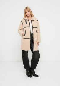 Cappotto beige in finto shearling con motivo a griglia a righe nere, cerniere e tasche laterali. Indossato sopra una maglietta chiara e jeans neri a vita alta.