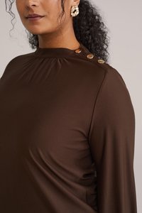 Blusa marrone con collo alto, arricciature sulle spalle e bottoni dorati su un lato; tessuto liscio e leggermente lucido.