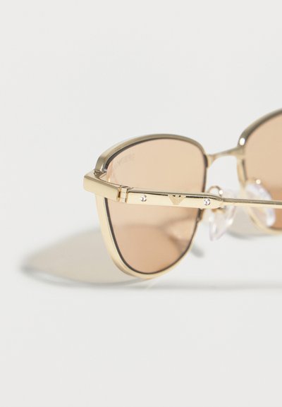 Γυαλιά ηλίου aviator με χρυσή κορνίζα, φακούς καφέ απόχρωσης και διακριτικό λογότυπο στον βραχίονα, τοποθετημένα πάνω σε ανοιχτόχρωμη επιφάνεια.