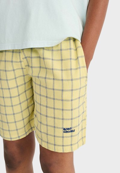 Personne portant un short jaune à carreaux avec des lignes de quadrillage bleues et un logo blanc sur la jambe gauche, associé à une chemise blanche.