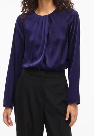 Femme portant un chemisier violet brillant à manches longues avec des plis au niveau du col, rentré dans un pantalon noir taille haute.