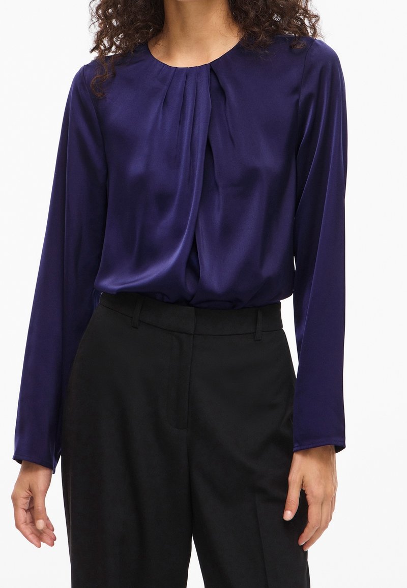 Femme portant un chemisier violet brillant à manches longues avec des plis au niveau du col, rentré dans un pantalon noir taille haute.