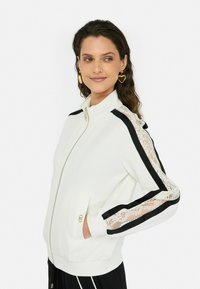 Veste zippée blanche avec des accents de rayures noires et des détails en dentelle sur les manches. Elle présente un col haut et des poches latérales. Quincaillerie dorée comprise.