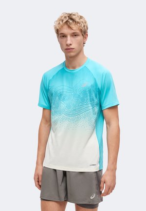 Junger Mann mit kurzen lockigen blonden Haaren, der ein hellblau-weiß gemustertes kurzärmeliges Sportshirt und graue Shorts trägt.