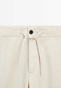 Gros plan sur la ceinture d'un pantalon en lin beige avec un bouton noir et un cordon en tissu noué pour un ajustement réglable.