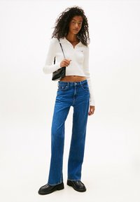 Mujer con cabello rizado que lleva una camiseta blanca de manga larga corta, jeans rectos azules, mocasines negros y lleva un pequeño bolso negro de hombro.