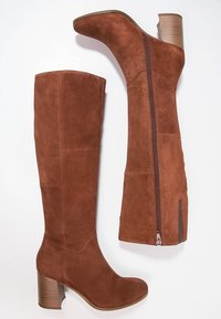 Paire de bottes marron en daim montant jusqu'au genou, avec talons blocs superposés et fermetures éclair latérales, présentées de profil et de dos sur un fond blanc.