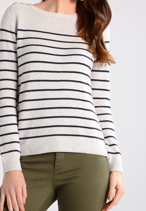 Kvinde iført en langærmet beige sweater med sorte vandrette striber og olivengrønne tætsiddende bukser mod en ensfarvet baggrund.