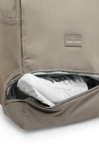 Beige hátizsák, alsó részén cipzáras zsebbel, benne egy fehér sneaker található, texturált talppal és fűzőkkel. A márkás címke látható.