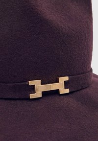 Fialový klobouk z filcu s širokým okrajem a zlatým kováním na pásku, s měkkou texturou a strukturovaným tvarem.