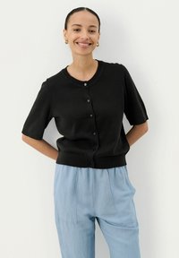 Schwarze gestrickte Kurzarm-Cardigan mit rundem Ausschnitt, Knopfverschluss und geripptem Saum, kombiniert mit hellblauen lockeren Hosen.