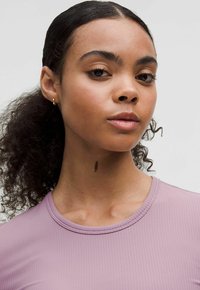 Top ribbed de culoare mov deschis, cu guler rotunjit, cu o textură netedă. Modelul are părul creț și poartă un cercei mic din aur în formă de inel.