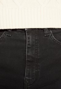 Jeans de mezclilla negra con un diseño ajustado, que presentan una textura suave, costuras visibles y una cintura alta. Un suéter de punto crema parcialmente visible.