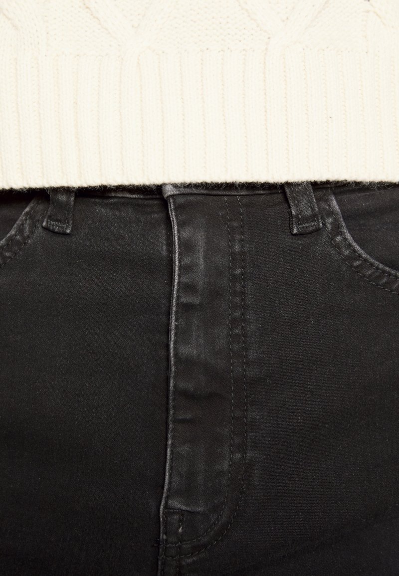 Jeans de mezclilla negra con un diseño ajustado, que presentan una textura suave, costuras visibles y una cintura alta. Un suéter de punto crema parcialmente visible.