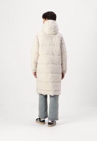 Manteau rembourré long beige clair avec une capuche, design à coutures verticales, poignets côtelés et ourlet arrondi, associé à un jean en denim et des sneakers.
