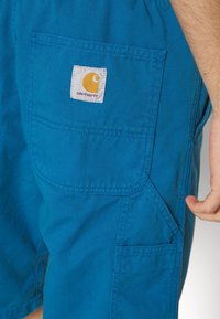 Niebieskie bawełniane szorty z tylną kieszenią z białą metką, na której znajduje się żółte logo oraz tekst "carhartt". Gładka tekstura, swobodny fason.