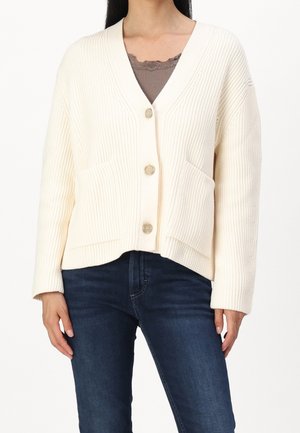 Cream textured cardigan with a V-neck, three buttons, and two front pockets. Worn over a brown lace top with blue jeans.