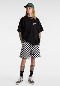 Sort oversized t-shirt, ternede sort-hvide shorts, grøn cap og brune sneakers. Har tatoveringer på synlige ben og mærkevarer som accessories.