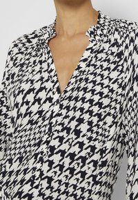 Blus med svart och vit houndstooth-mönster som har en texturerad tyg, en krage och knappdetaljer fram.
