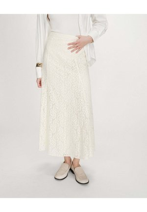 Femme portant une longue jupe en dentelle blanche, un haut côtelé blanc, une chemise blanche et des mocassins beiges, debout devant un fond uni.