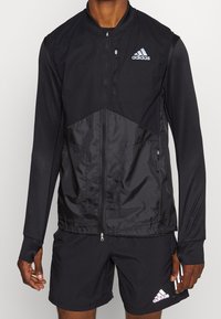 Black athletic jacket with a zip front, featuring textured panels and a logo. Long sleeves and elastic cuffs, paired with black shorts.