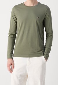 Oliwkowy, długi rękaw T-shirt z miękkiego, lekkiego materiału, z okrągłym dekoltem i małym logo po lewej stronie na piersi, w zestawieniu z białymi spodniami.