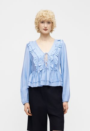 Γυναίκα με κοντά σγουρά ξανθά μαλλιά φορά ανοιχτό μπλε μπλούζα με μακριά μανίκια και βολάν, καθώς και μαύρο σορτς μέχρι το γόνατο, στέκεται μπροστά σε λευκό φόντο.