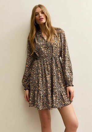 Młoda kobieta o długich blond włosach, ubrana w mini sukienkę z długim rękawem, w kwiatowy wzór, z taliowaniem, na jednolitym beżowym tle.